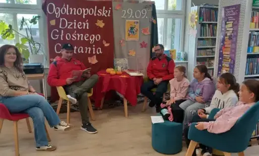 Na zjęciu czwórka dzieci z Dyskusyjnego Klubu Książki, moderatorka klubu oraz dwoje zaproszonych gości - strażacy z OSP Dobieżyn czytajacy dzieciom. W tle dekoracja z okazji Ogólnopolskiego Dnia Głośnego Czytania.